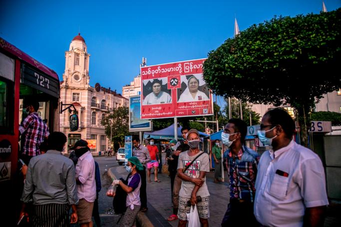 People wearing facemasks wait for a bus under an election campaign poster in Yangon on October 7, 2020. Photo: Sai Aung Main / AFP